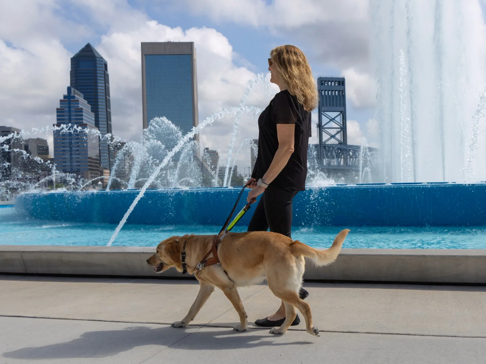 A woman using a guide dog in harness to navigate near a fountain in downtown Jacksonville