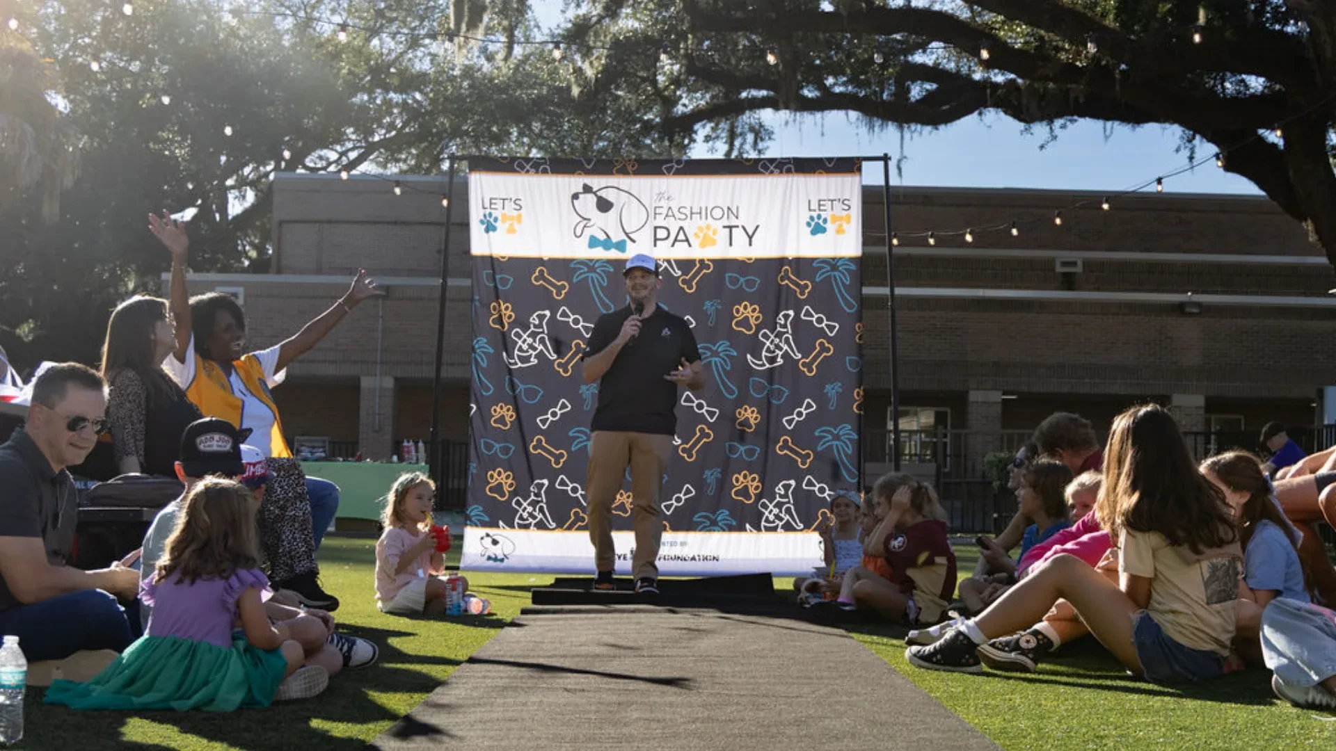 Emcee on stage at The Fashion PAWty 2026 runway at Happy Brew, Jacksonville, with families and children seated on the lawn