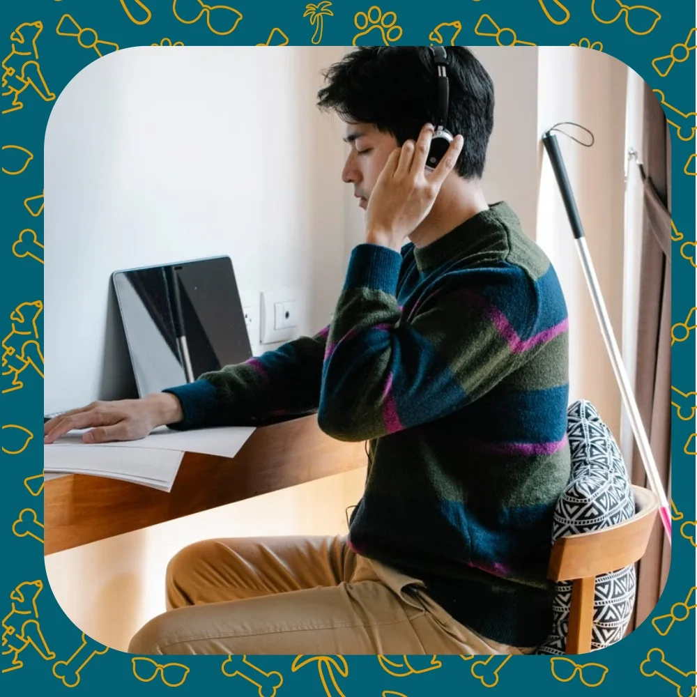 A man wearing headphones works at a desk with a white cane resting nearby
